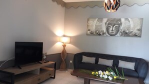 Una sala de estar con un televisor sobre un soporte de madera, un sofá, una mesa de centro con flores, una lámpara y una obra de arte en la pared.