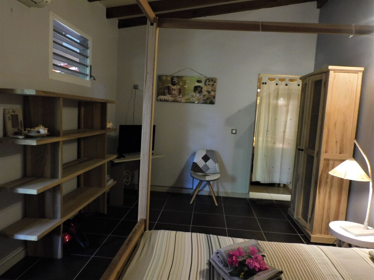 A room with a wooden floor, a bed with a striped bedspread, a chair, a shelf unit, a painting on the wall, and a window with blinds.