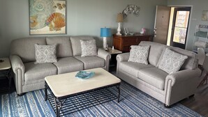 A living room with two grey sofas, a coffee table, a patterned rug, a painting, and a wooden cabinet. - Fantastic beachfront 2-bedroom condo with AC in pleasant Tybee Island (Tybee Island)