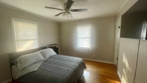Quarto com cama, ventilador de teto e duas janelas com persianas.