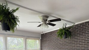 Quarto com parede de tijolos, ventilador de teto, mesa com cadeiras e plantas penduradas.