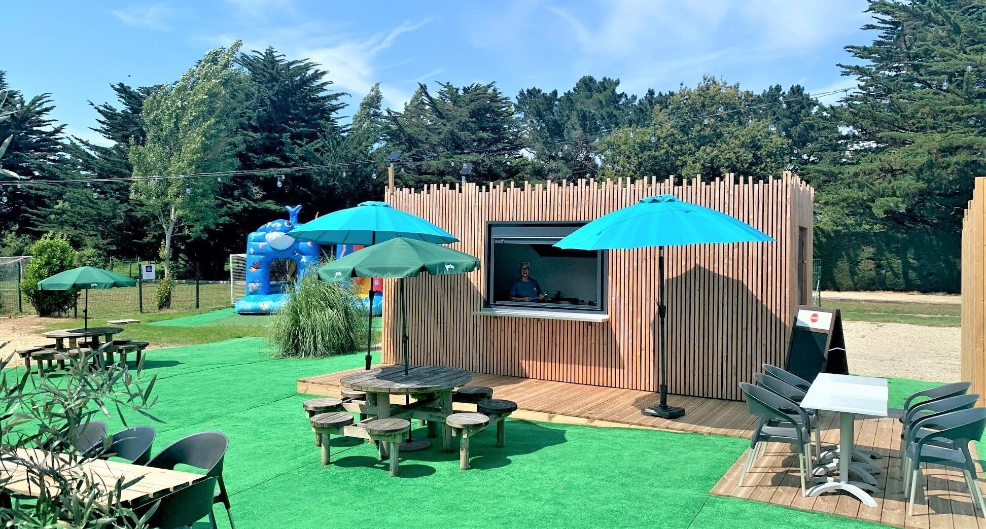 Eine Außenpatio-Terrasse mit Holztischen und -stühlen, eine kleine Holzhütte mit Fenster und eine blaue aufblasbare Rutsche im Hintergrund.