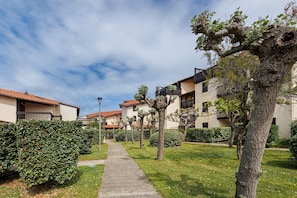 Un chemin mène à une rangée de bâtiments dotés de balcons, le tout étant entouré de haies et d’arbres bien entretenus.