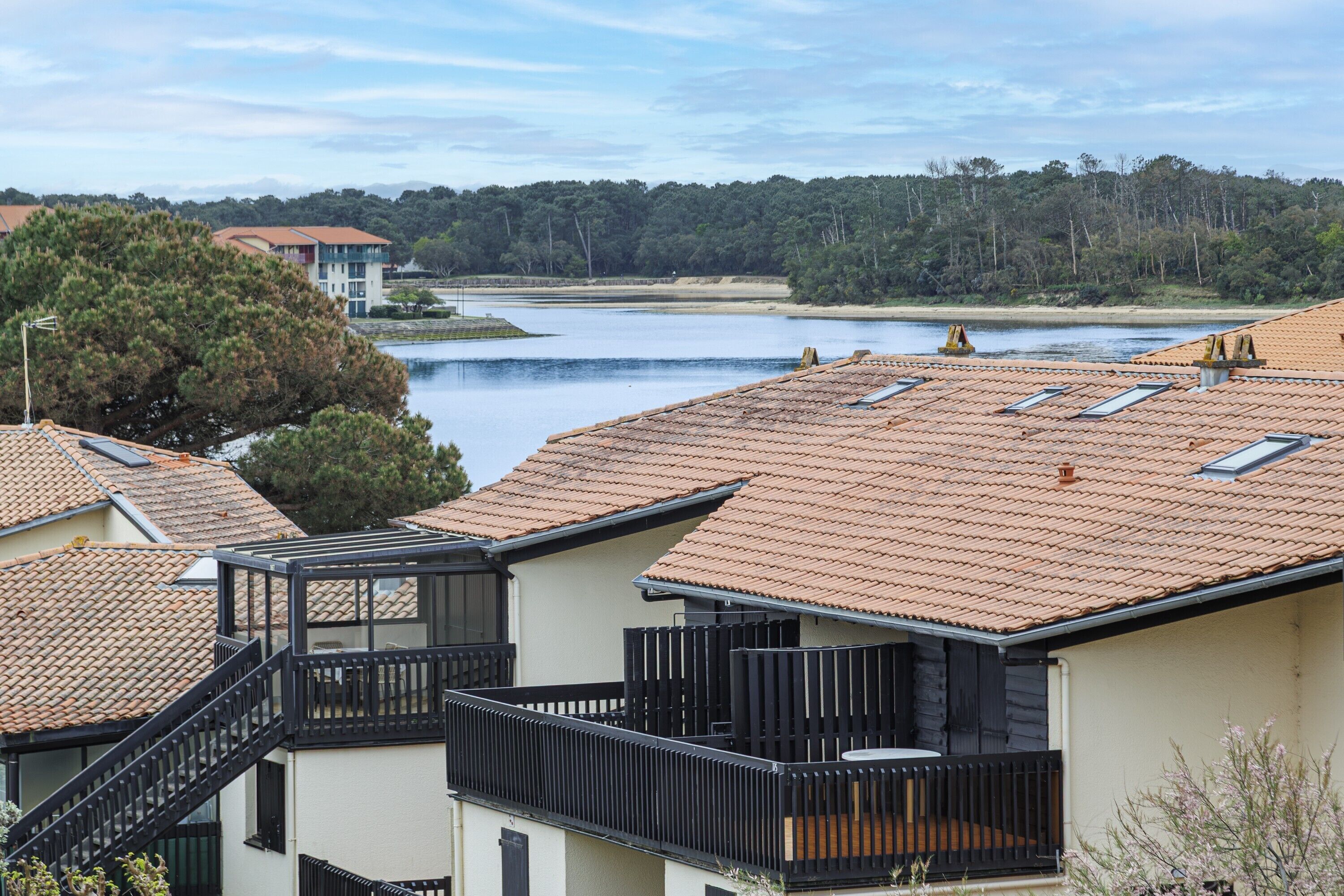 Un barrio residencial con tejados de tejas color terracota, un balcón con barandilla negra y vistas a un lago y colinas boscosas al fondo.