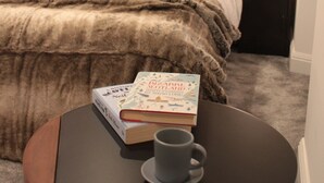 Ein Schlafzimmer mit einer gepolsterten Kopfteile, einem Bett mit einer braunen Decke und zwei Kissen, einem Nachttisch mit einem Buch und einer Tasse sowie Holzvertäfelung an der Wand.