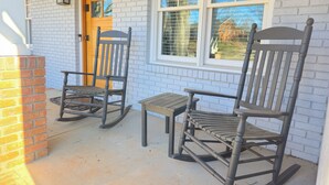 A porch with two rocking chairs and a small table, a brick wall, and a window with blinds. - Downtown Mooresville 3BR Brick Home - Fully Renovated (Mooresville)