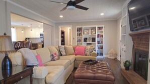 A living room with a sectional sofa, a leather ottoman, a bookshelf, a fireplace, and a ceiling fan. - Downtown Mooresville 3BR Brick Home - Fully Renovated (Mooresville)