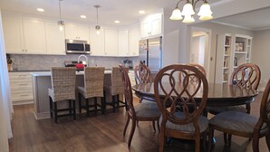 A modern kitchen with white cabinets, a stainless steel refrigerator, and a dining area with wooden chairs and a dark wooden table. - Downtown Mooresville 3BR Brick Home - Fully Renovated (Mooresville)