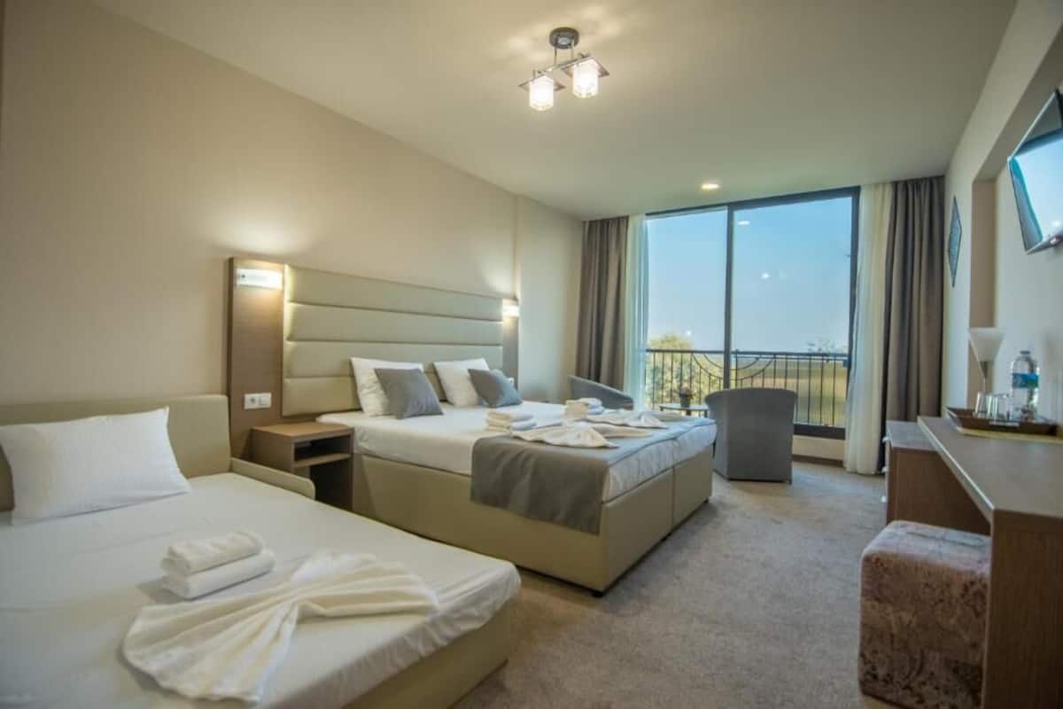 A hotel room with two beds, a desk, a chair, and a balcony with a view.