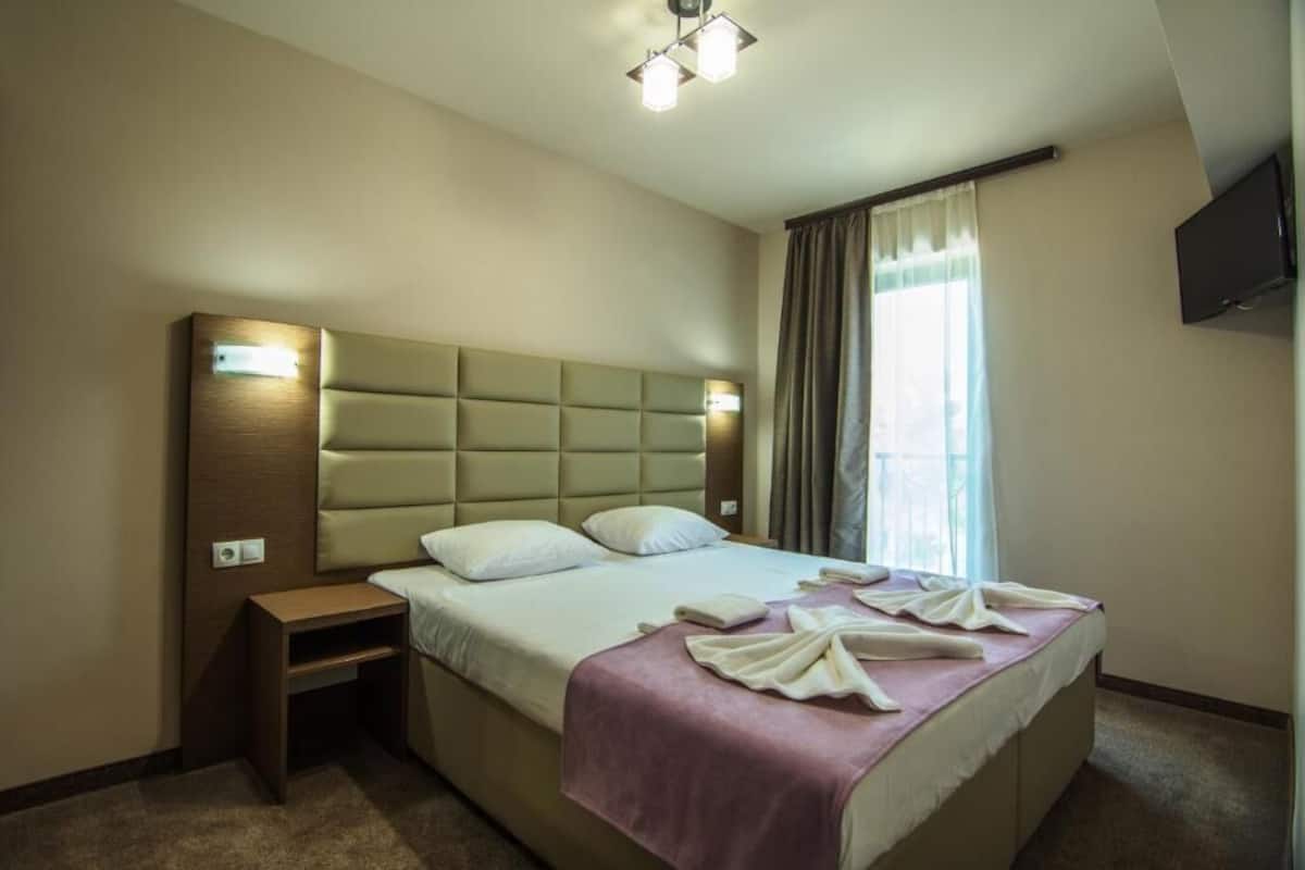 A hotel room with a large bed, a nightstand, a flat-screen TV, and a window with curtains.