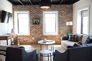 A living room with a brick wall, two windows, a round table, and a sofa.