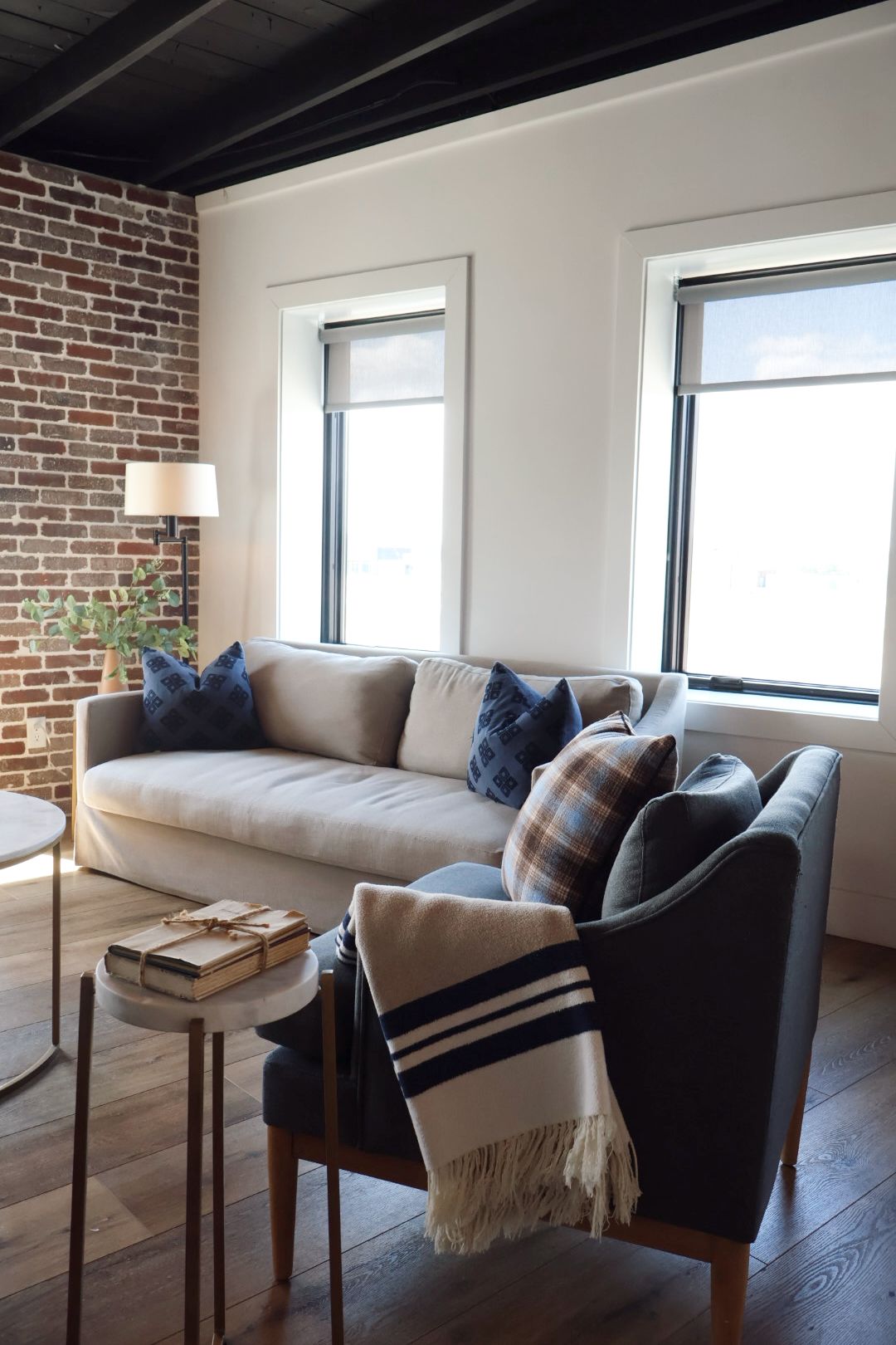 A living room with a sofa, a chair, a small table, and a lamp. There are windows and a brick wall.