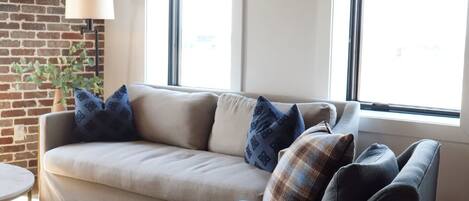 A living room with a sofa, a chair, a small table, and a lamp. There are windows and a brick wall.