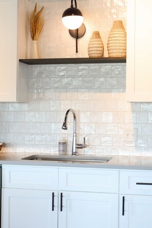 A kitchen with white cabinets, a stainless steel sink, a black pendant light, and woven vases on a shelf.