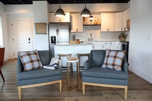 Ein modernes Wohnzimmer mit zwei grauen Sofas, einem kleinen Couchtisch und einer Küche im Hintergrund mit weißen Schränken und einem Edelstahlkühlschrank.