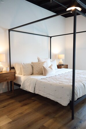 A four-poster bed with a white bedspread and multiple pillows, a wooden nightstand with a lamp, and a wooden floor.