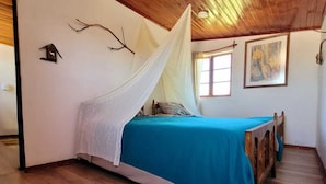 Une chambre avec un plafond en bois, un lit recouvert d’une courtepointe bleue, un baldaquin blanc et un petit nichoir fixé au mur.
