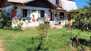 Une maison de style rustique, avec une façade blanche et des peintures murales florales aux couleurs vives, un porche en bois et un jardin au premier plan.