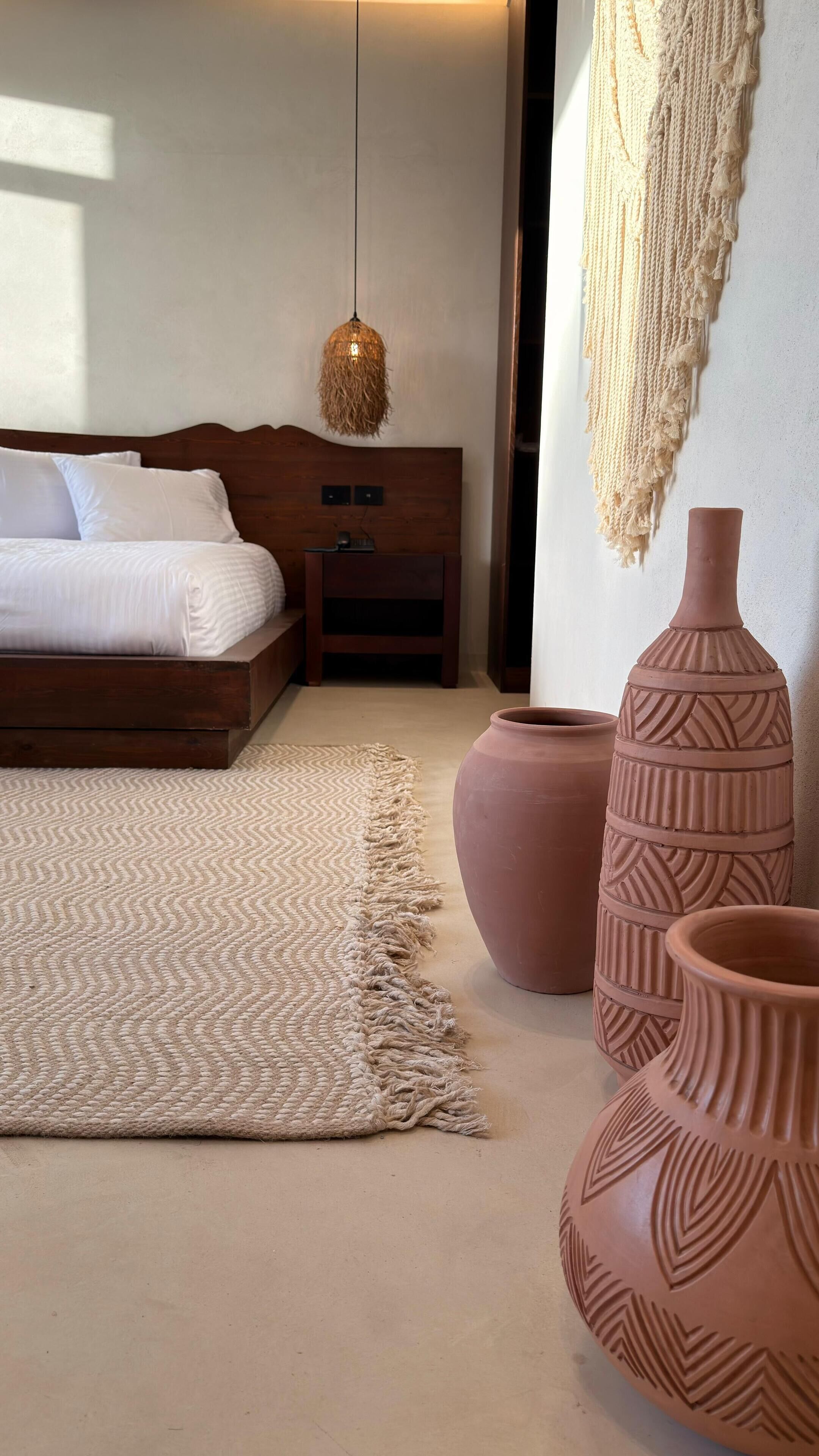 A bedroom with a wooden bed, a woven rug, terracotta pots, and a macrame wall hanging.