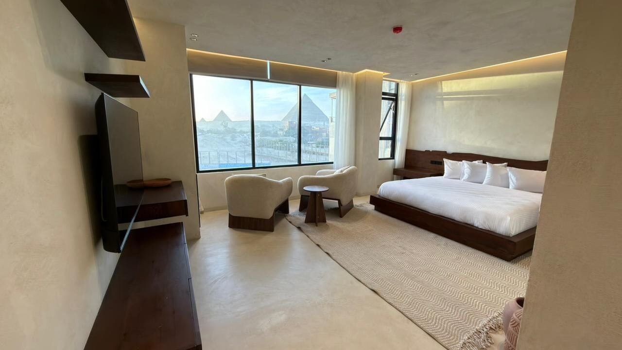 Een moderne hotelkamer met een groot bed, een zithoek met twee stoelen, een kleine tafel en uitzicht op een piramide door grote ramen.