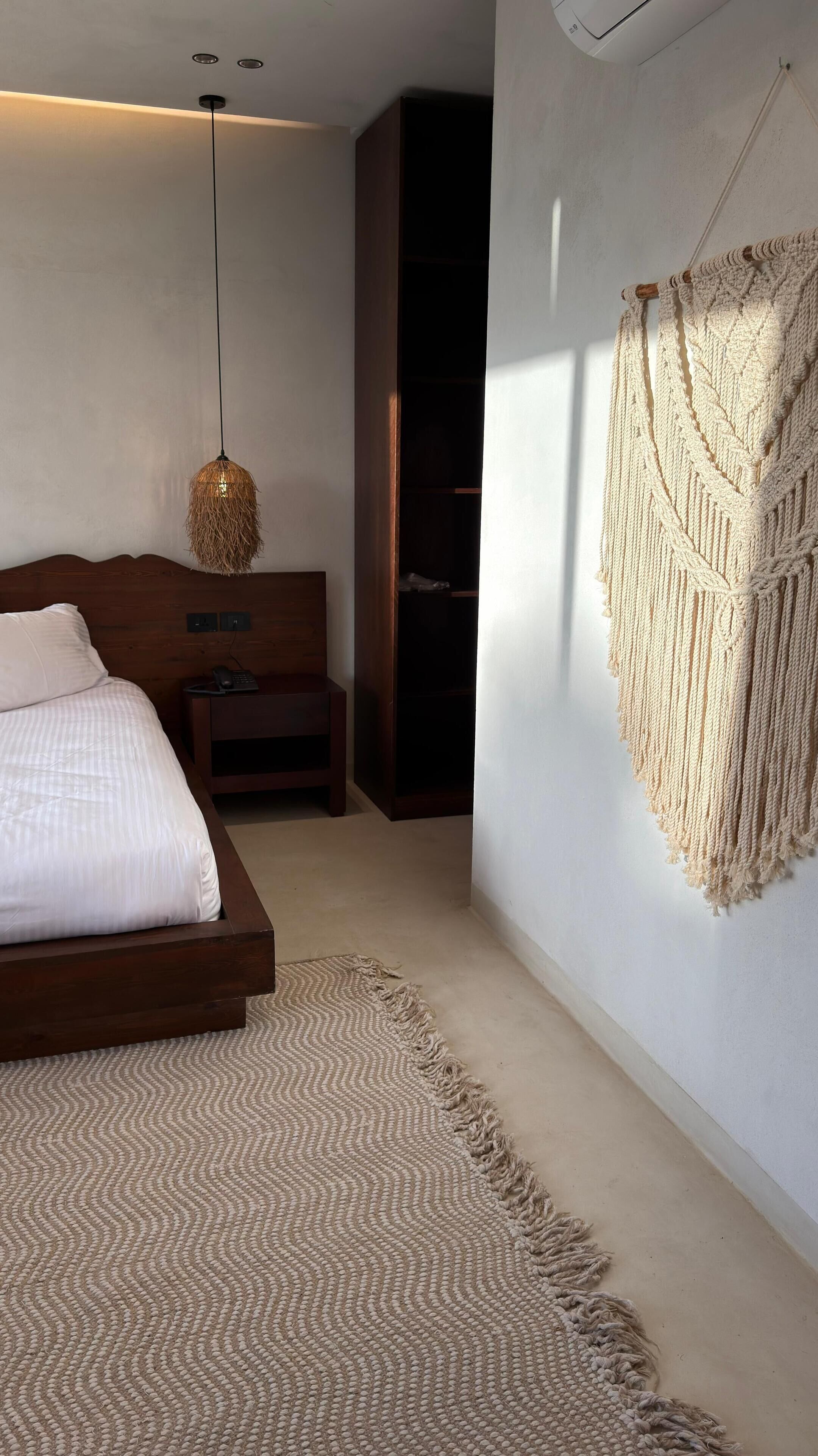 A bedroom with a bed, bedside table, hanging macrame wall decor, and a woven rug.