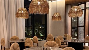 Un espace repas avec des tables rondes et des chaises en osier, de grandes fenêtres et des lustres suspendus en osier.