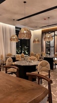 A dining area with round tables and wicker chairs, large windows, and hanging wicker chandeliers.