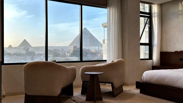 Une chambre d’hôtel dotée d’une grande fenêtre donnant sur les pyramides. On y trouve deux fauteuils et une petite table.