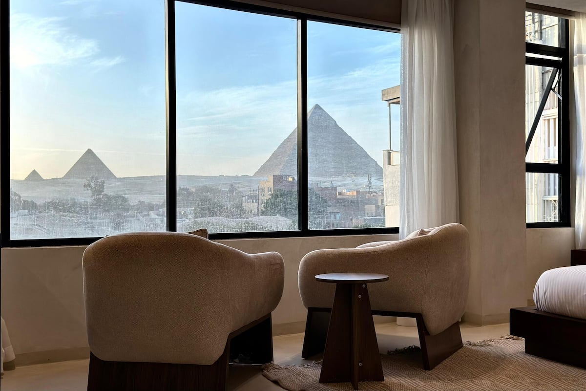A hotel room with a large window offering a view of the pyramids. There are two armchairs and a small table.