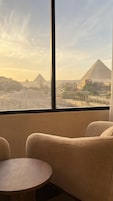 A room with a large window overlooking a cityscape and pyramids. There are two chairs and a small round table.
