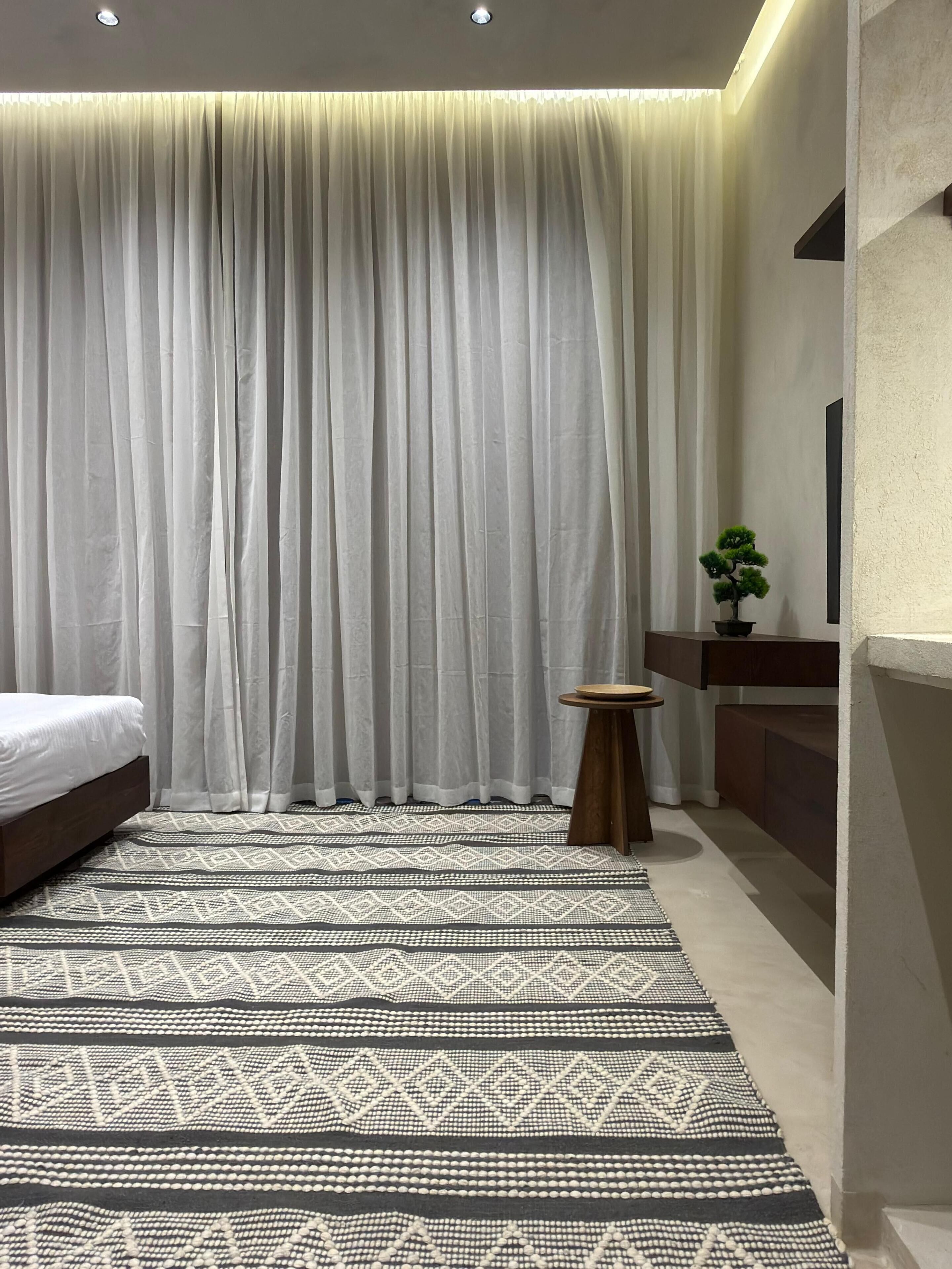 A modern bedroom with a bed, a patterned rug, a small round table, a potted plant, and a large window with curtains.