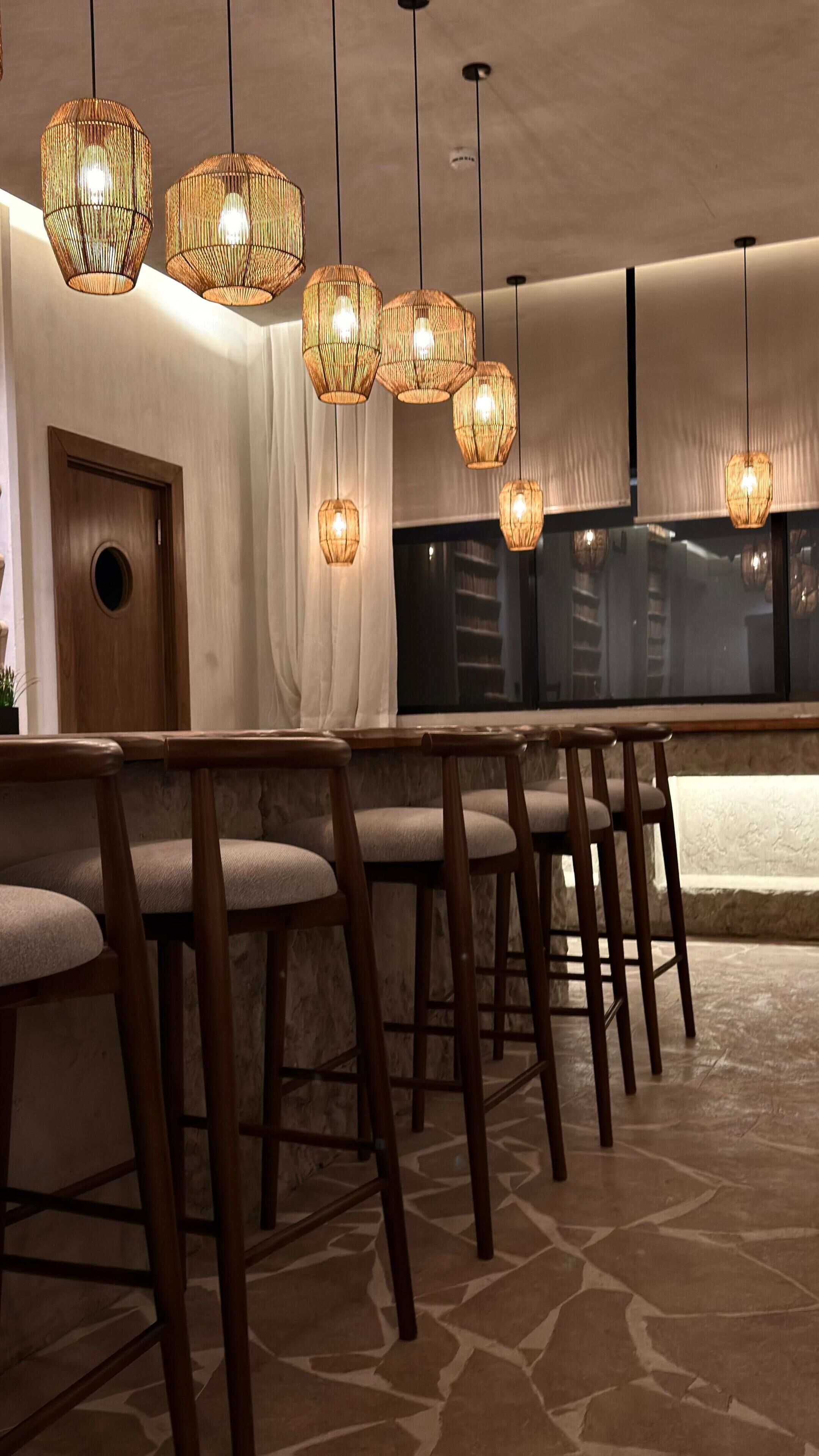 Un ristorante con tavoli e sedie in legno, lampadari in vimini appesi e una vista sulla città attraverso ampie finestre.