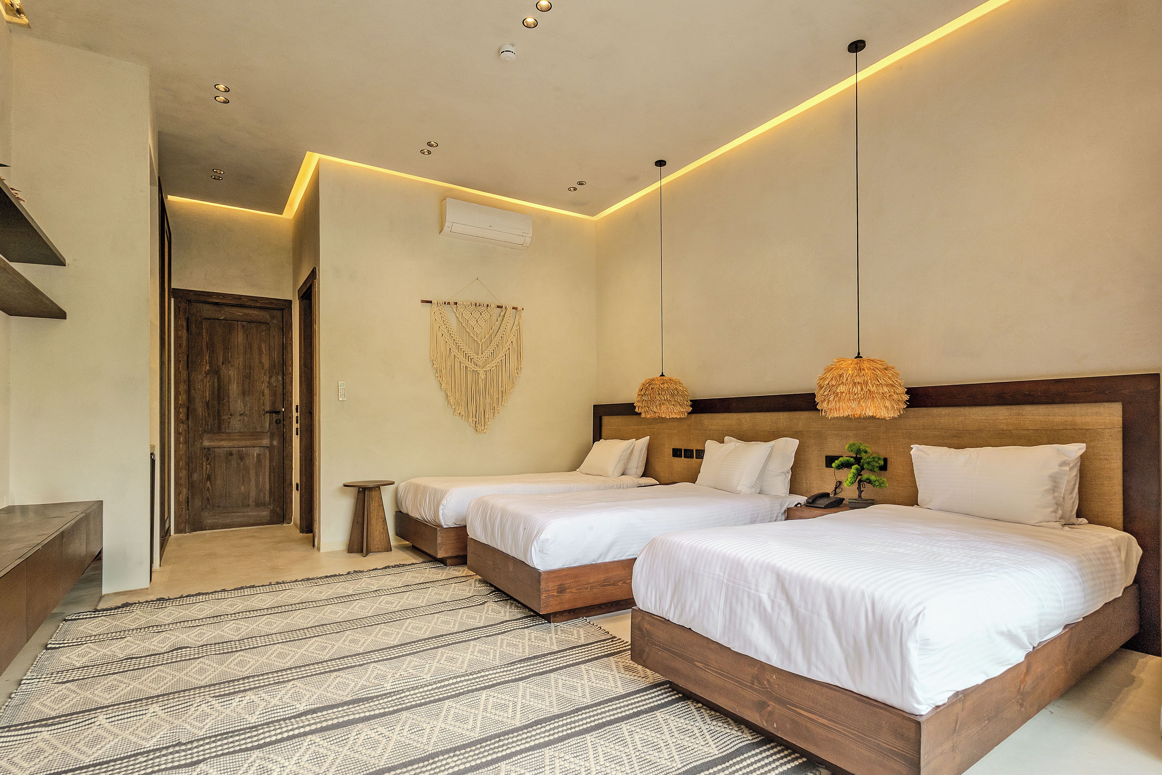 A hotel room with two beds, a wooden headboard, a hanging macrame wall decoration, and a patterned rug.