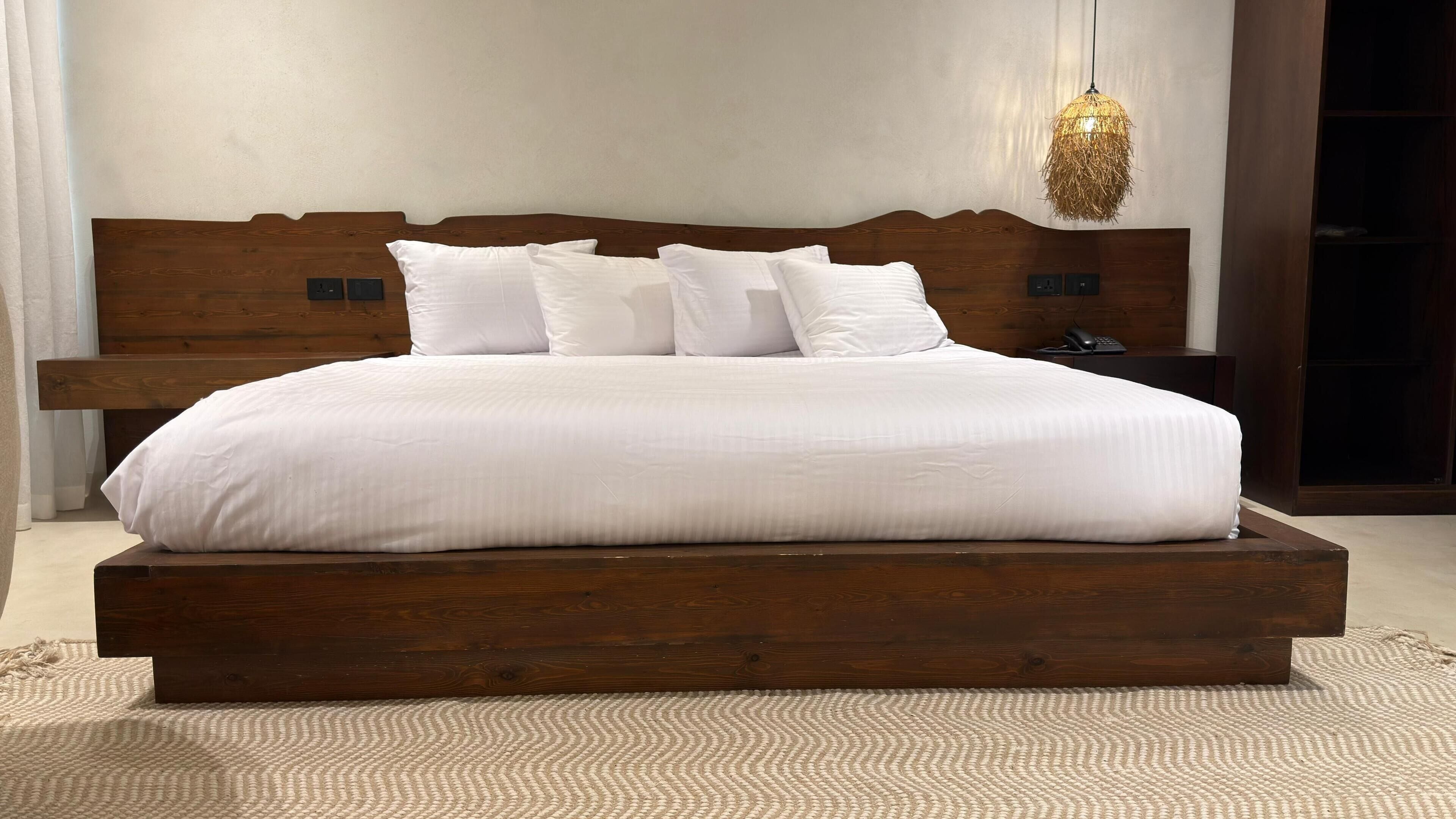 A large bed with a wooden frame and white bedding, a woven light fixture above, and a nightstand with a phone.