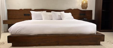 A large bed with a wooden frame and white bedding, a woven light fixture above, and a nightstand with a phone.