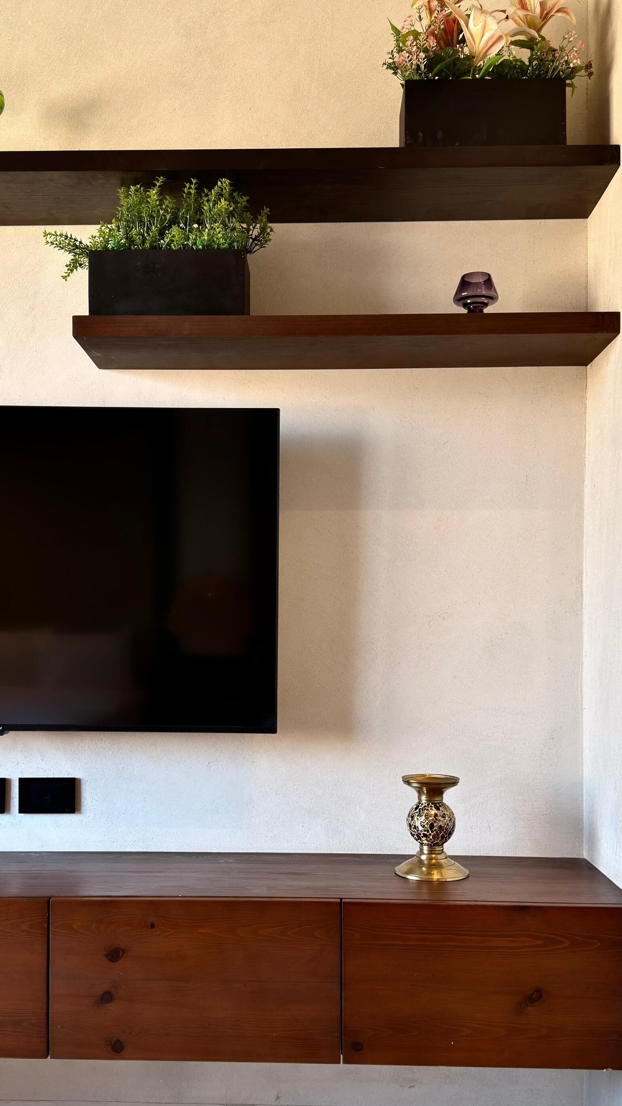 Een moderne woonkamer met een flatscreen televisie aan de muur, houten planken met decoratieve objecten, en een kleine tafel met een messing kandelaar.