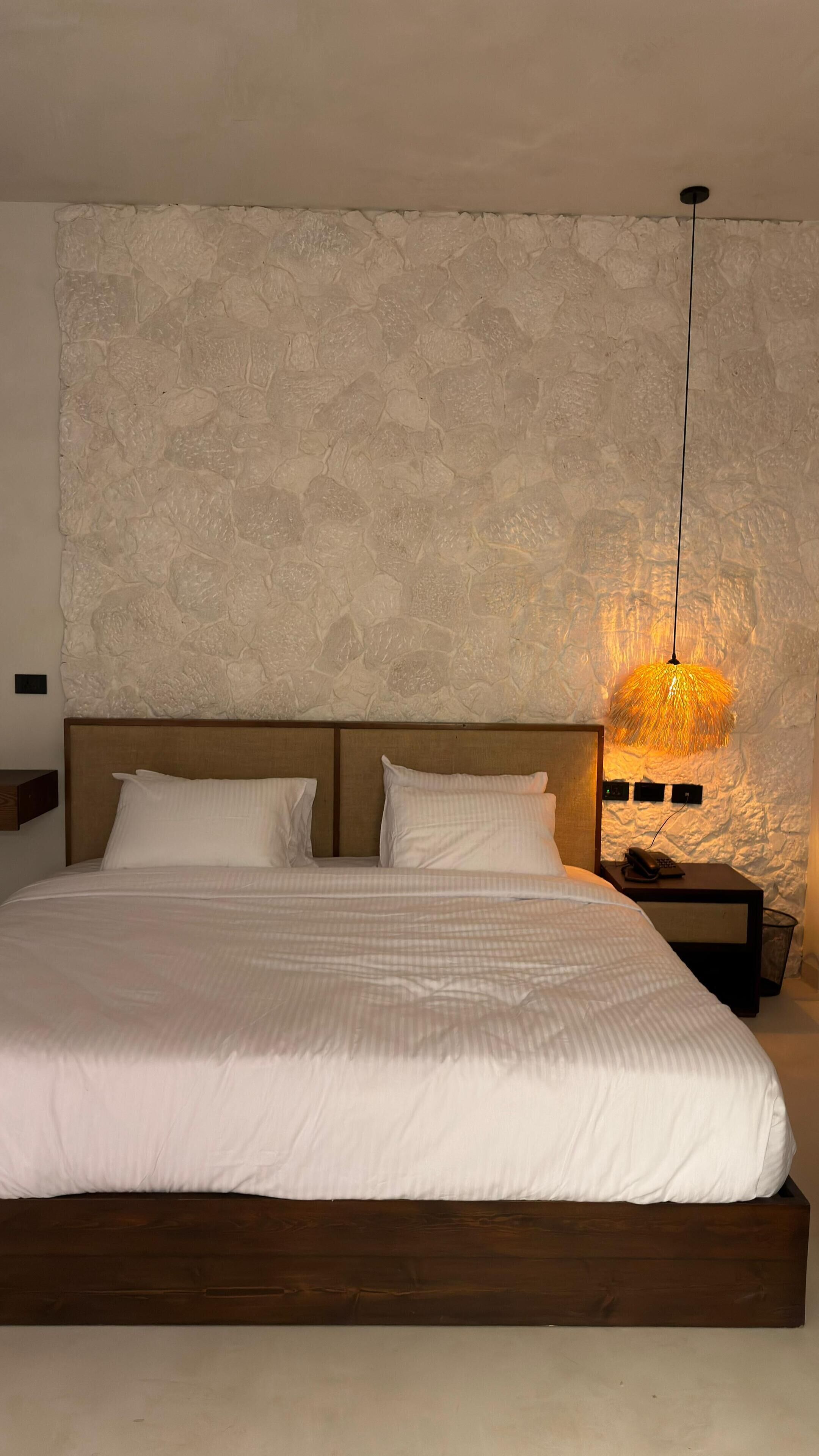 A bedroom with a large bed, a textured wall, a hanging lamp, and a nightstand with a phone.