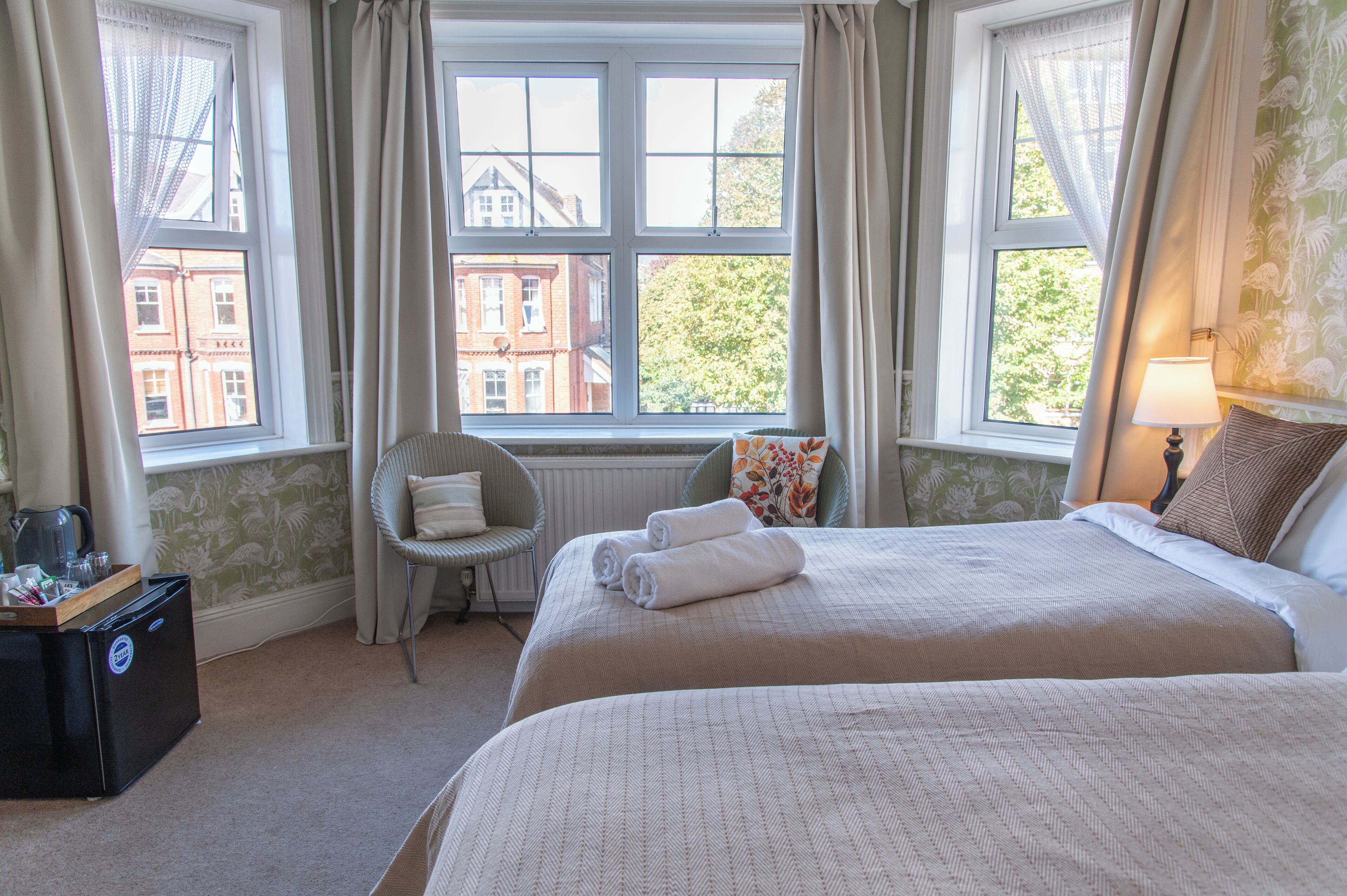 A hotel room with two beds, a chair, a radiator, and a view of buildings through large windows.