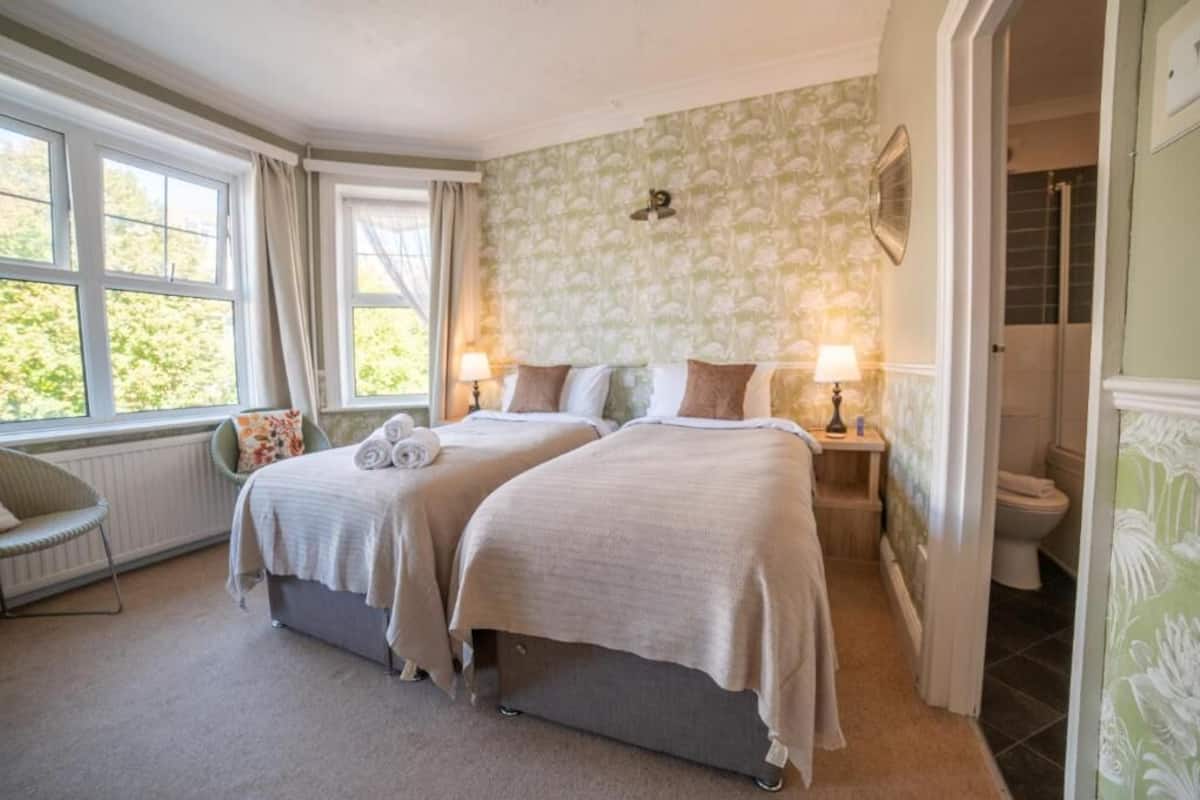 A bedroom with two beds, a window with curtains, a chair, and a bathroom visible through an open door.