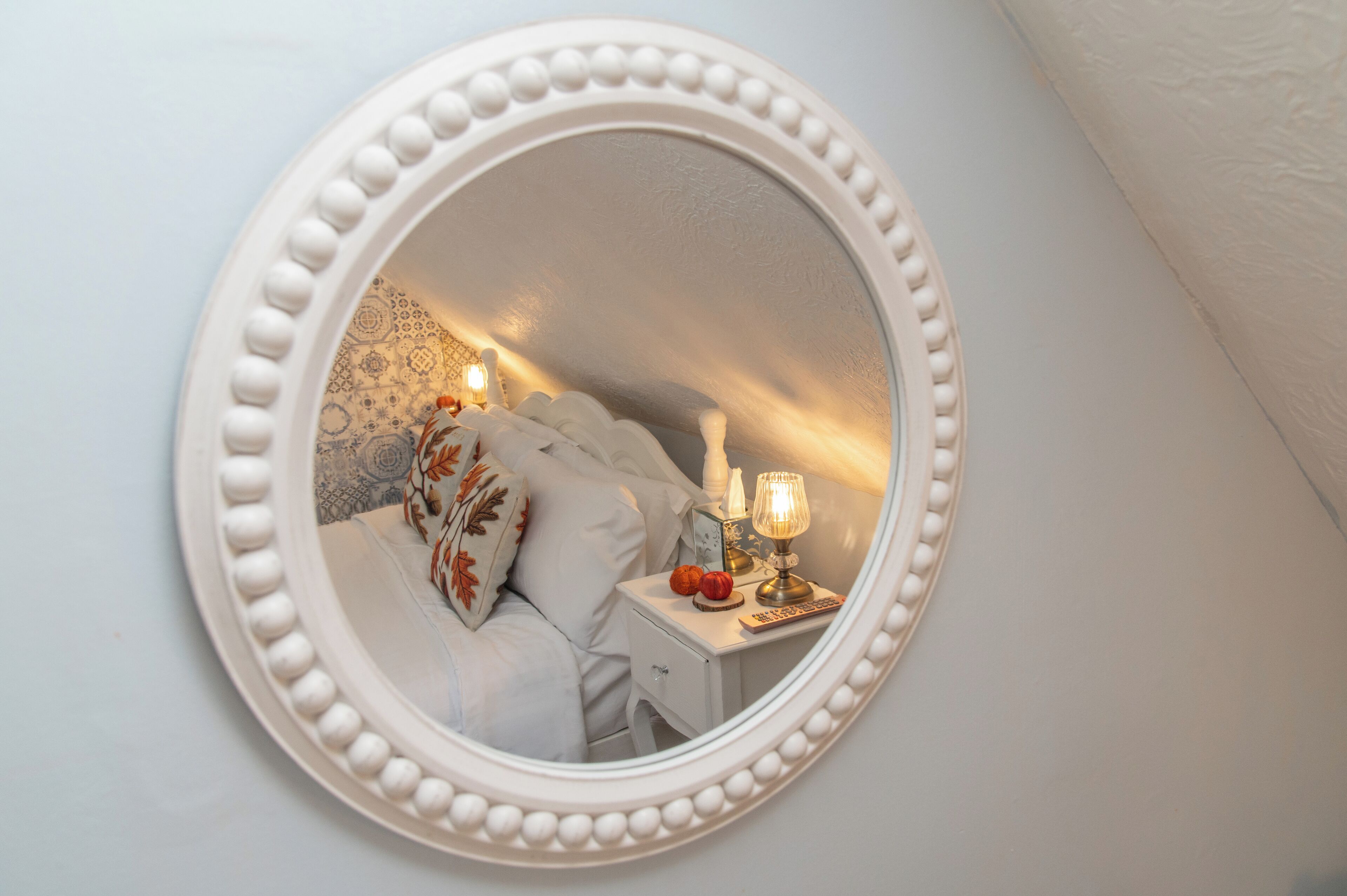 A bedroom with a round mirror, a bed with white bedding, a nightstand with a lamp, and a decorative wall.