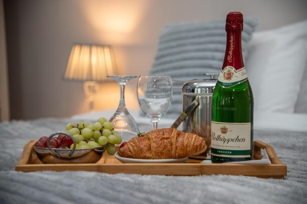A tray with a bottle of Rölkäppchen, grapes, a croissant, and glasses on a bed.