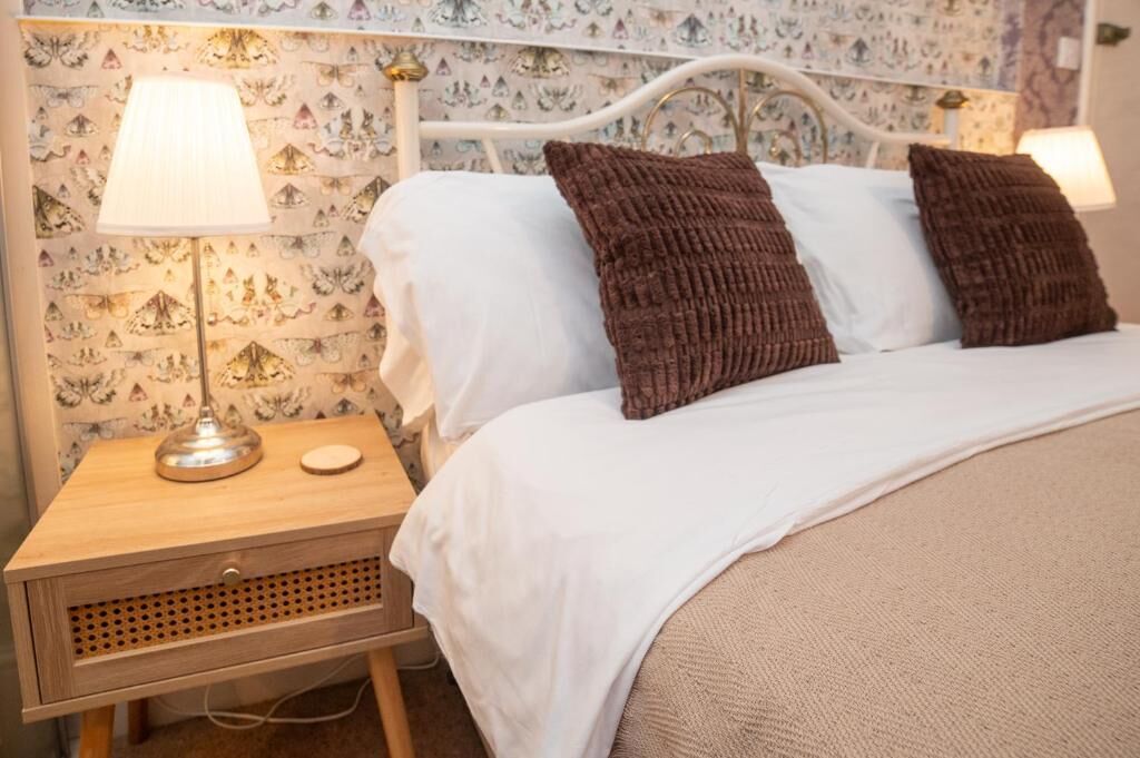 Una cama bien hecha con almohadas blancas y marrones, una mesita de noche de madera con una lámpara, y un papel tapiz estampado.