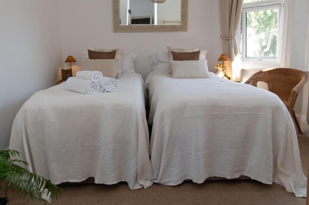Une chambre d’hôtel avec deux lits simples, un grand miroir, une fenêtre avec des rideaux, un fauteuil en osier et une petite table avec une lampe.