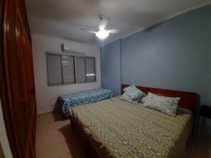 A bedroom with two beds, a ceiling fan, a window with blinds, and a wooden door. - Pitangueiras Center, ocean view, air conditioning, 3 bedrooms, balcony, 1 parking space, barbecue. (Guarujá)