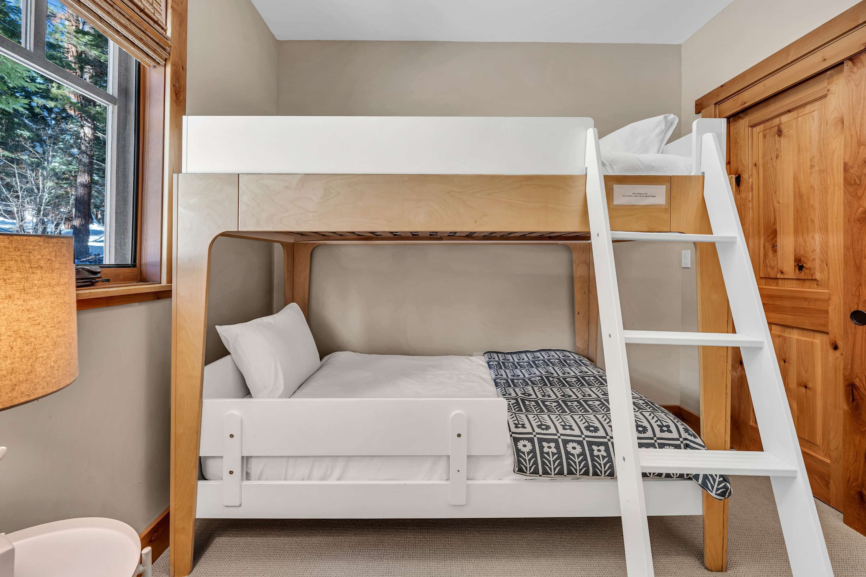 Een stapelbed met een ladder, een raam met uitzicht op bomen, en een houten garderobe.