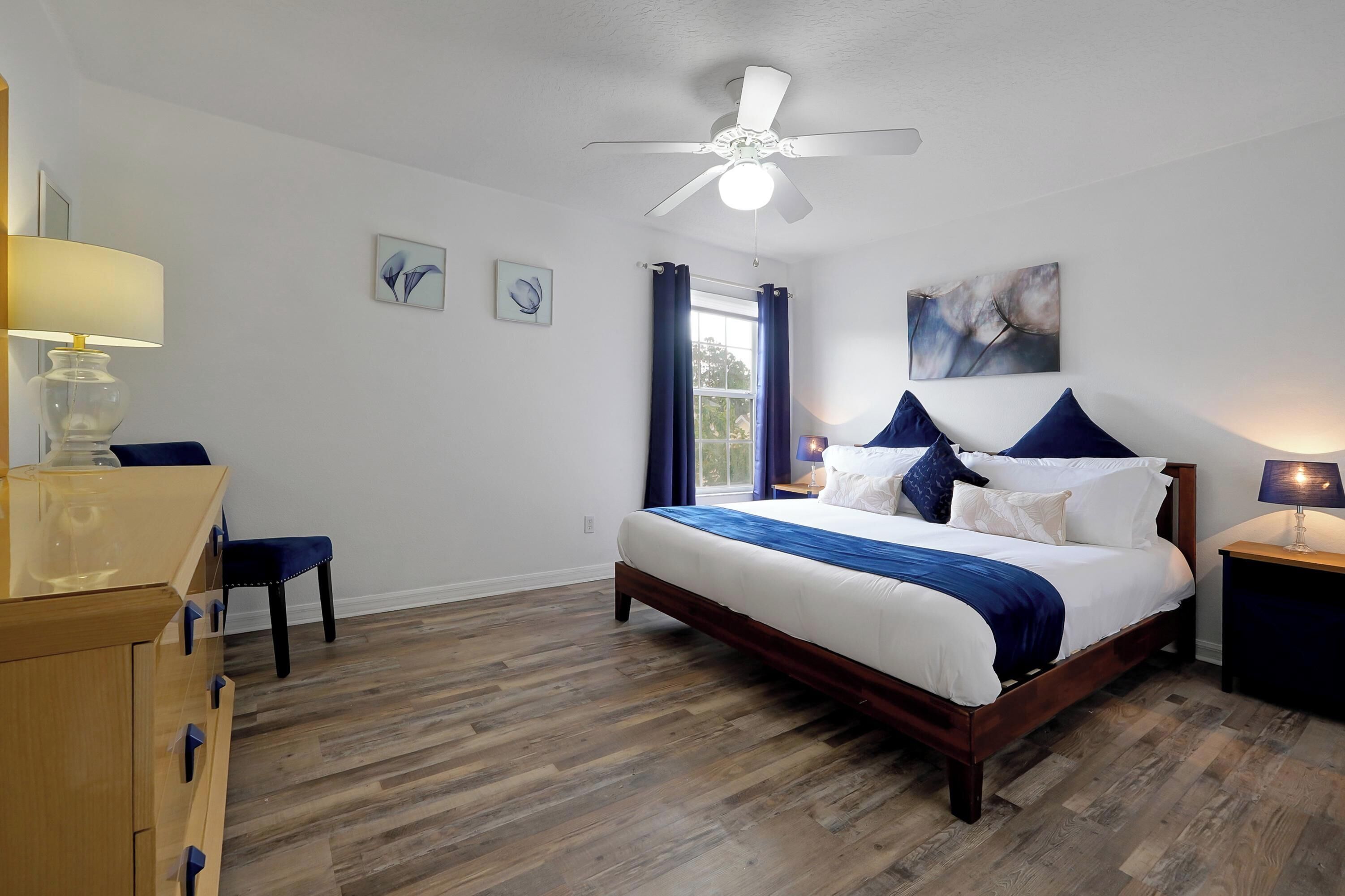 A bedroom with a wooden bed, a ceiling fan, a desk with a lamp, and a window with curtains.