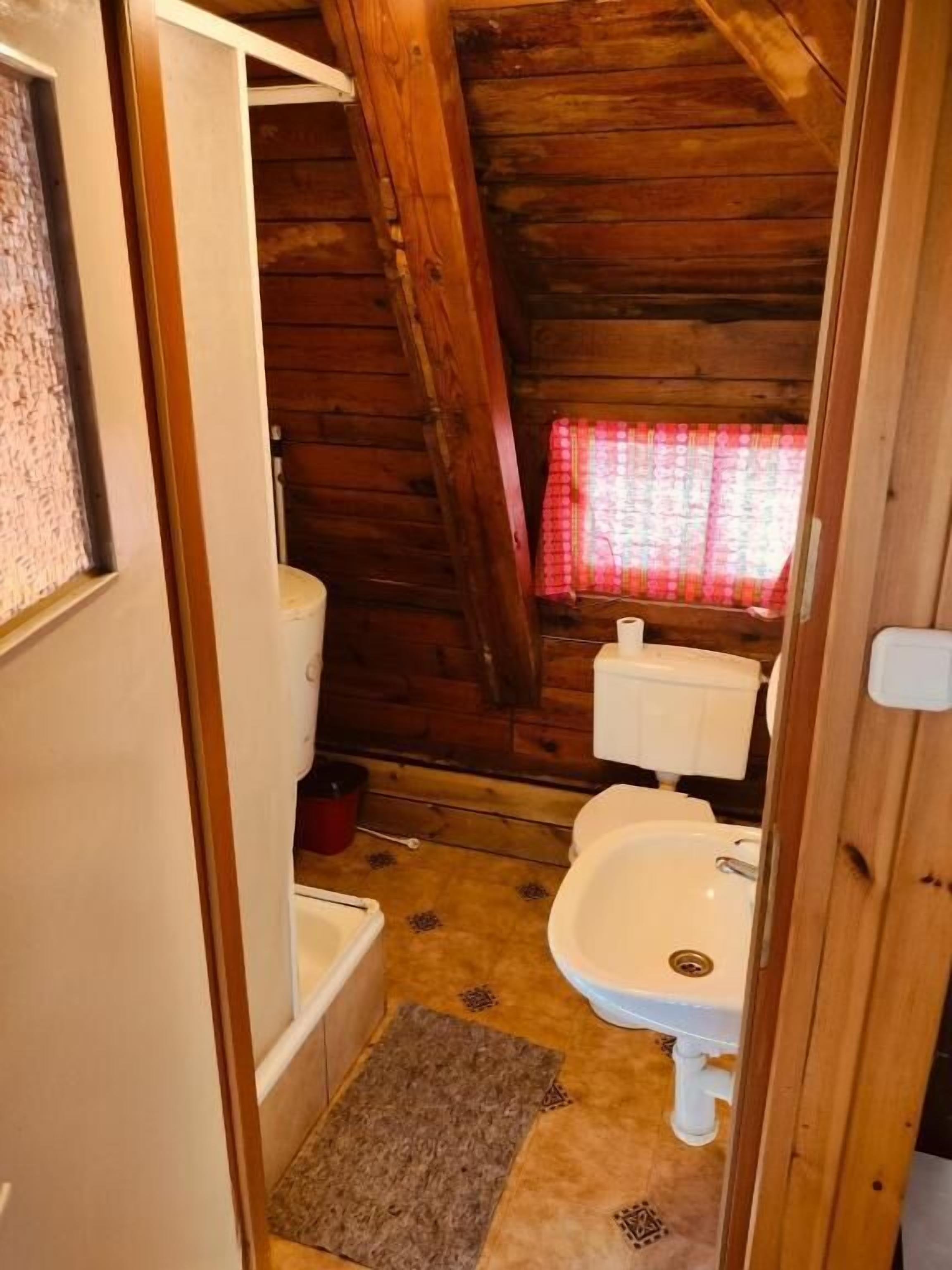 Kapea, ullakkokerroksessa sijaitseva kylpyhuone, jonka seinät ovat puuta. Siellä on wc-istuin, pesuallas ja ikkuna, jossa on punainen verho.