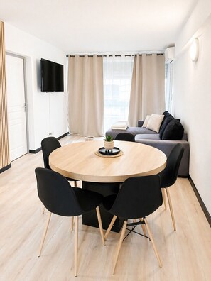 A modern living room with a round wooden dining table, black chairs, a grey sofa, and a flat-screen TV mounted on the wall. - LVAM - Appartement T3 - 6 personnes - Au coeur du Cap d'Agde - Piscine (Agde)