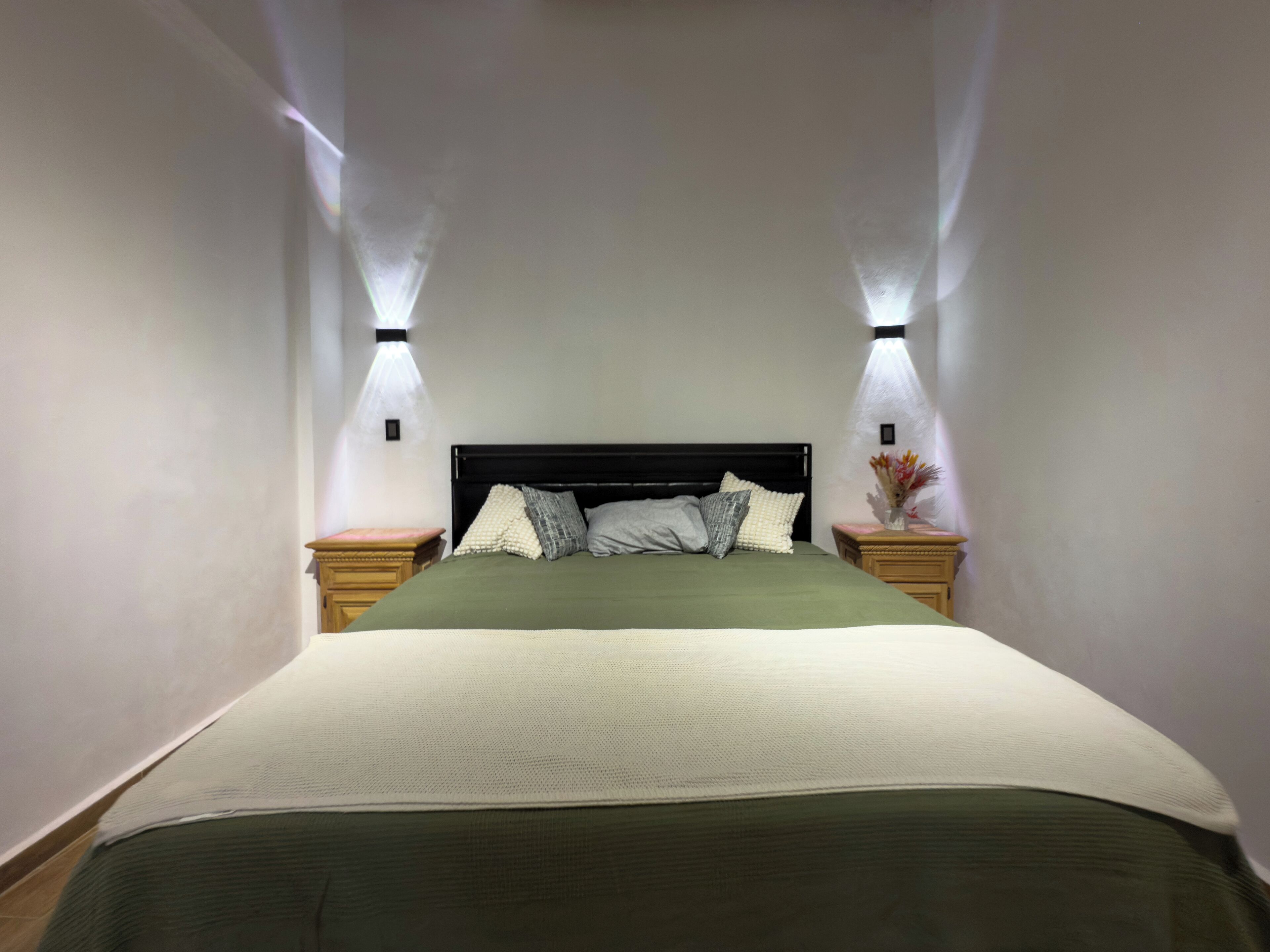 A neatly made bed with a green bedspread and multiple pillows, flanked by two bedside tables with lamps, in a room with plain walls and a simple, modern design.