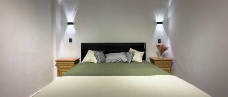 A neatly made bed with a green bedspread and multiple pillows, flanked by two bedside tables with lamps, in a room with plain walls and a simple, modern design.